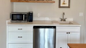 Cocina compacta con muebles blancos, lavavajillas de acero inoxidable, microondas y cafetera. Hay estantes de madera sobre la encimera con vajilla y un cuadro enmarcado.