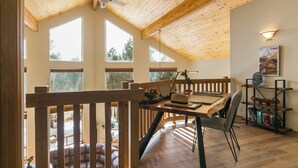 Ruime kamer met een hoge plafond, houten balken, grote ramen, een eethoek met tafel en stoelen, en een boekenkast.
