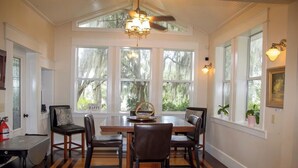 A dining room with a large wooden table, six chairs, and a ceiling fan. There are large windows with views of trees, and a fire extinguisher is visible on the left.