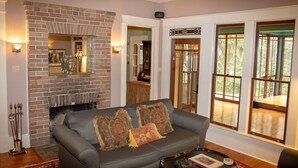 A living room with a brick fireplace, a grey sofa with patterned cushions, and large windows offering a view of the outdoors.
