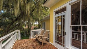 A wooden deck with two chairs and a table, a yellow building with large windows, and trees with hanging moss.
