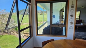 Ein Zimmer mit einem großen Fenster, das Blick auf ein Wiesfeld und Bäume bietet. Es gibt einen Holztisch mit Stühlen sowie einen kleinen Tisch mit einer Pflanze.