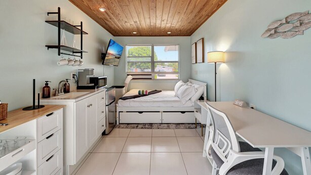 A compact bedroom with a bed, bedside table, and a desk. There is a window with a view of greenery outside. - Coastal Comfort 4BR in Stuart FL (Stuart)
