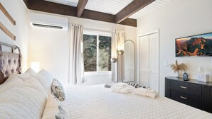 Una cama grande con cabecera, una mesita de noche con lámpara, una ventana con cortinas y un televisor fijado a la pared.
