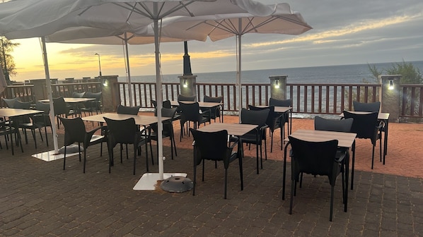 An outdoor patio with tables and chairs, umbrellas, and a view of the sea.