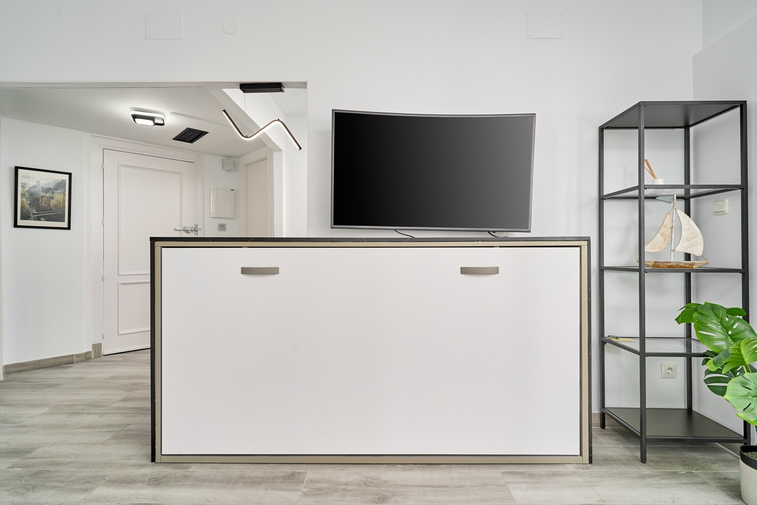 Un salón moderno con un televisor de pantalla plana montado en la pared, un mueble de entretenimiento blanco minimalista y una estantería metálica negra con una planta en maceta.