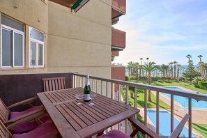 Ein Holztisch mit einer Flasche und Gläsern, ein Balkon mit Blick auf einen Pool und ein Gebäude mit Fenstern und Balkonen.