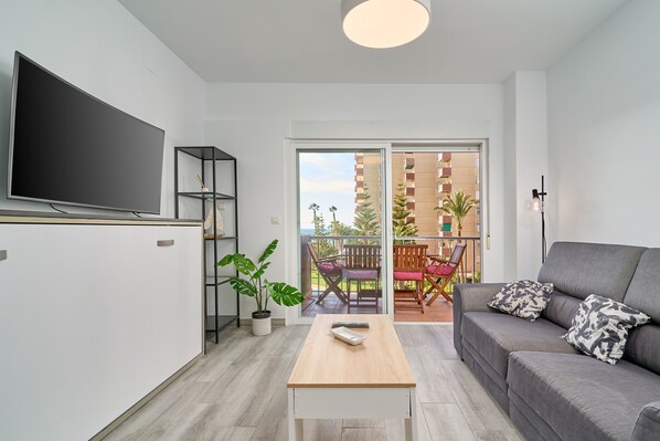 A modern living room with a flat-screen TV, a sofa, a coffee table, and a balcony with outdoor furniture.