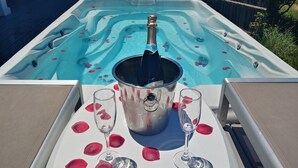 A hot tub with rose petals on the surface, a champagne bottle, and two glasses on a table.