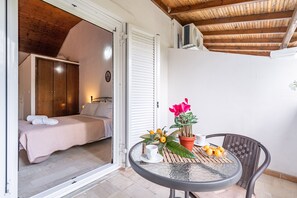 An open sliding door leading to a bedroom with a bed and a wardrobe, a round table with a potted plant and fruit, and a ceiling fan. - Studio Apartment 'Theodoros' with Shared Garden, Wi-Fi and Air Conditioning (Sidari)