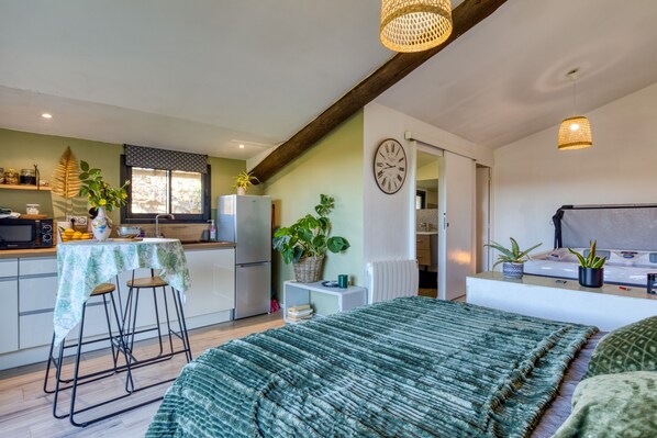 A compact kitchen with a dining area, a bed with a green blanket, and a clock on the wall.