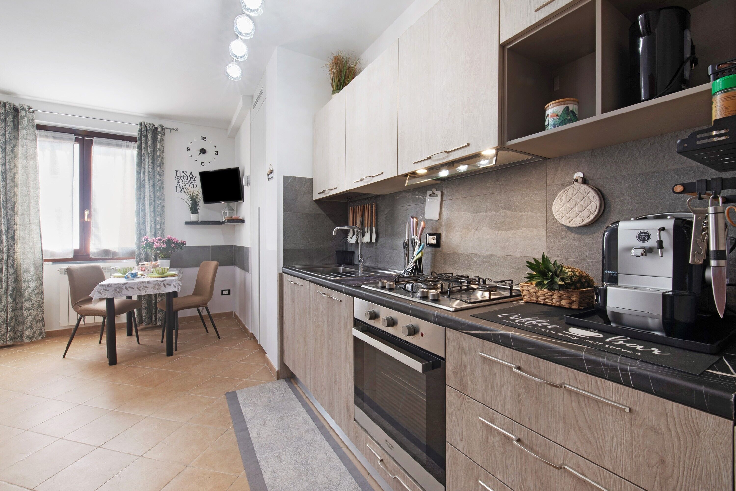Une cuisine moderne équipée d’une cuisinière à gaz, d’un four et d’une machine à café, un coin repas avec une table et des chaises, et un téléviseur fixé au mur.