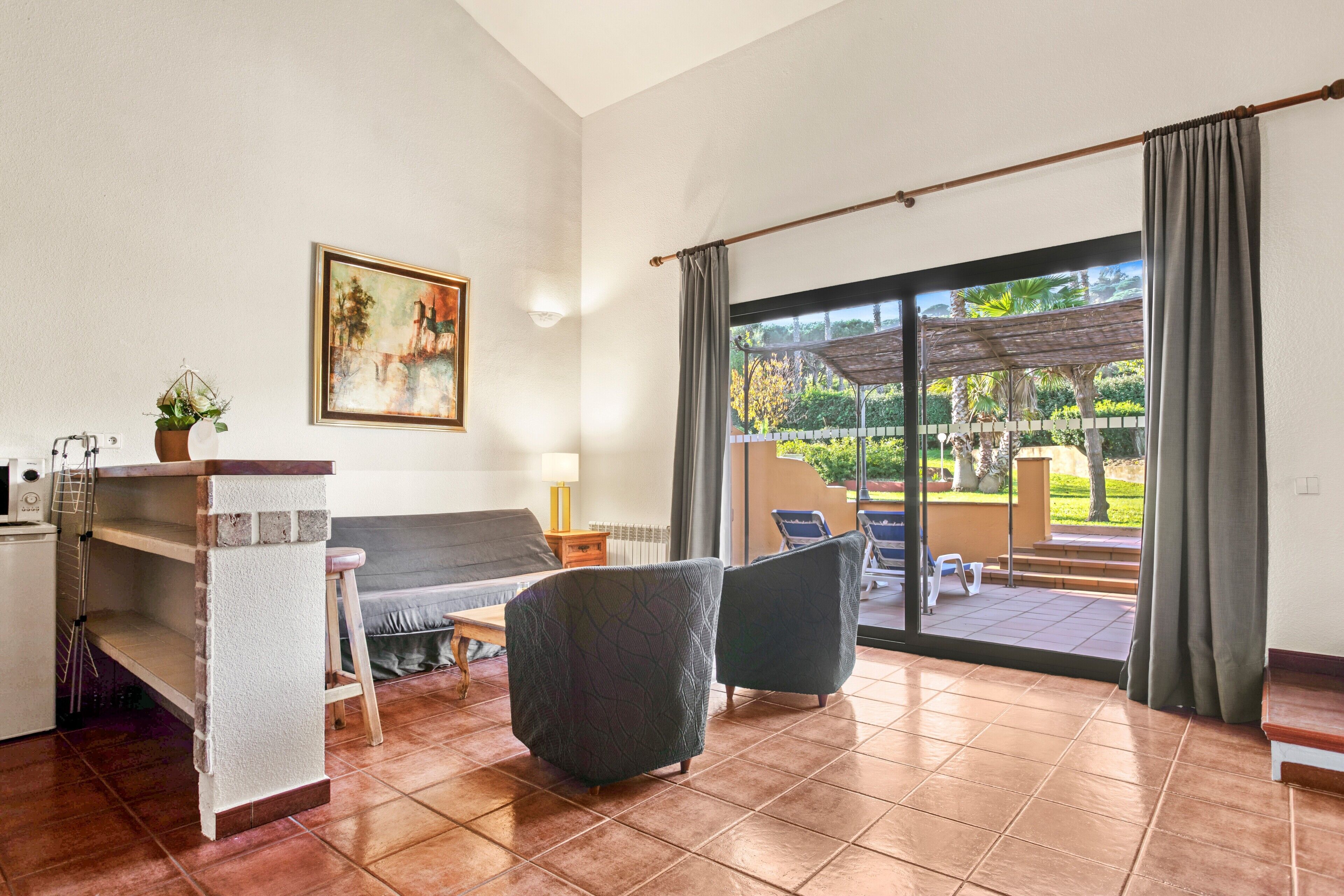 A living room with a sofa, two armchairs, a coffee table, a TV, a painting, and a view of an outdoor patio with a pergola.