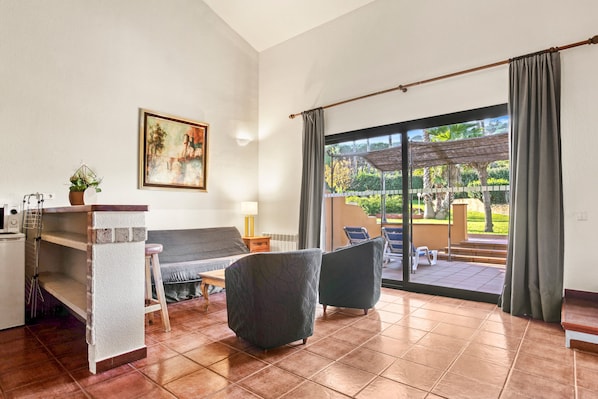 Sala de estar com sofá, duas poltronas, mesa de centro, televisão, quadro e vista para um pátio exterior com pérgola.