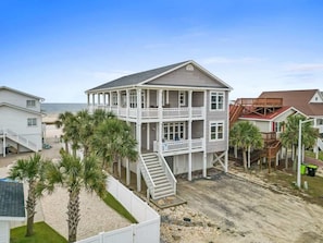A two-story beachfront house with a balcony, a staircase, and palm trees. - On the Rocks by Brunswick Vacation Rentals: 4BR OIB Retreat w/ Pool & Views (Ocean Isle Beach)