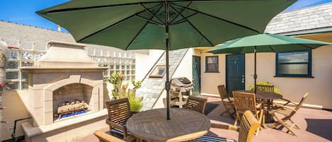 Un patio al aire libre con una mesa y sillas de madera redondas, una sombrilla verde, una chimenea de piedra y una parrilla.