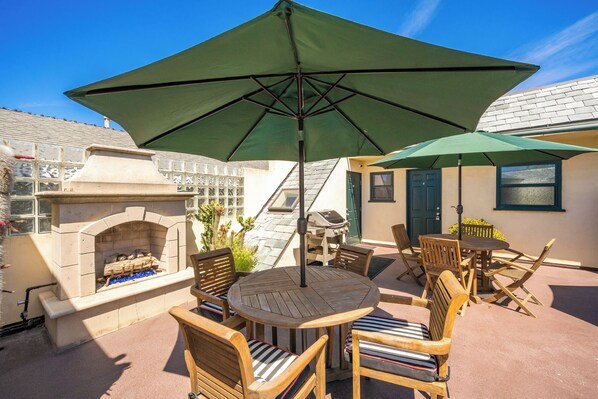 An outdoor patio with a round wooden table and chairs, a green umbrella, a stone fireplace, and a grill.
