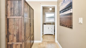 A hallway with a wooden wardrobe, a bathroom with a sink and mirror, and a beach scene painting on the wall.