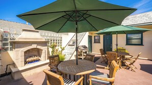 An outdoor patio with a round wooden table and chairs, a green umbrella, a stone fireplace, and a grill. - Clean Budget Friendly Apartment Steps from Beach (Newport Beach)