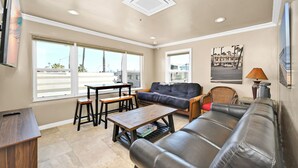A living room with a sofa, a coffee table, and a dining table. There are large windows and a ceiling vent. - Clean Budget Friendly Apartment Steps from Beach (Newport Beach)