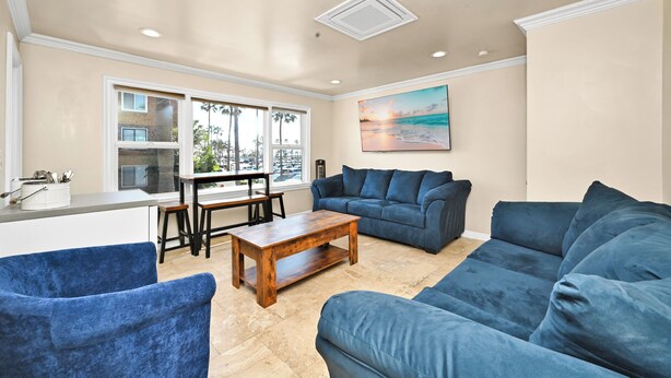 A living room with a beige ceiling, a large window with a view of palm trees, a blue sofa, a wooden coffee table, and a painting of a beach scene. - Partial Ocean View next to Boardwalk & Balboa Pier (Newport Beach)