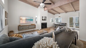 A spacious living room with a large gray sofa, a wooden coffee table, and a ceiling fan. There is a fireplace, a dining area with a kitchen in the background, and large windows providing natural light.