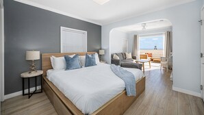 A hotel room with a large bed, a nightstand with a lamp, a seating area with a sofa and chairs, and a view of the beach. - West Oceanfront Oasis 1-2-3 (Newport Beach)