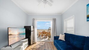 A modern living room with a blue sofa, a flat-screen TV, and a view of the beach through an open door. - West Oceanfront Oasis 1-2-3 (Newport Beach)