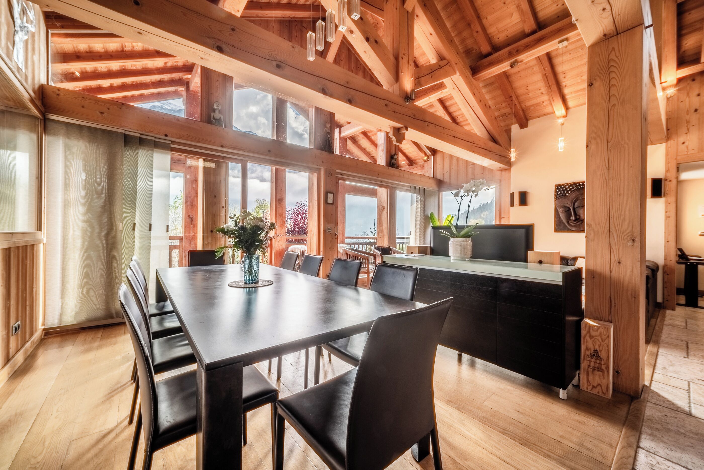 Une salle à manger spacieuse avec une longue table et des chaises, des poutres en bois au plafond et de grandes fenêtres offrant une vue dégagée.