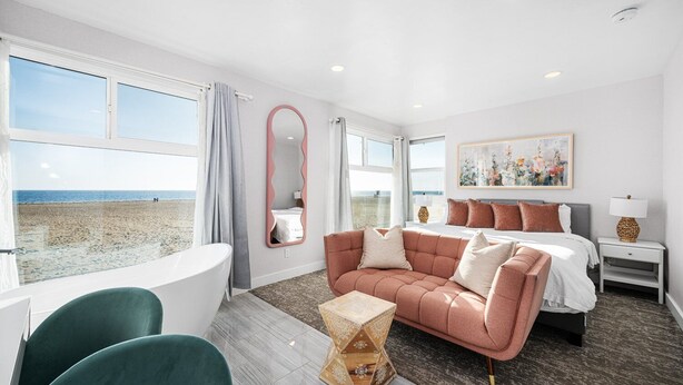 A hotel room with a large window overlooking a beach, a bed with multiple pillows, a green armchair, a small table, and a bedside lamp.