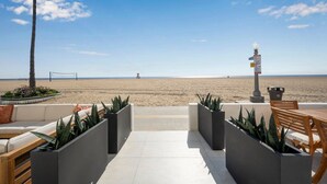 Une terrasse au bord de la plage avec vue sur l’océan, un filet de volley-ball et, au loin, une tour de surveillance des sauveteurs.