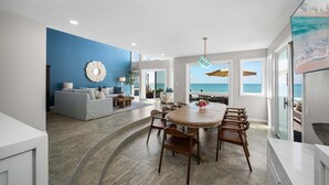 A modern living room with a dining area, large windows, and a view of the ocean. - Oceanfront Family Home with Firepit on the Sand (Capistrano Beach)