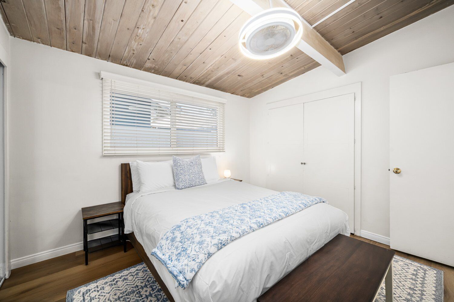 A bedroom with a wooden ceiling, a bed with white bedding and a blue floral throw, a bedside table, and a window with blinds.