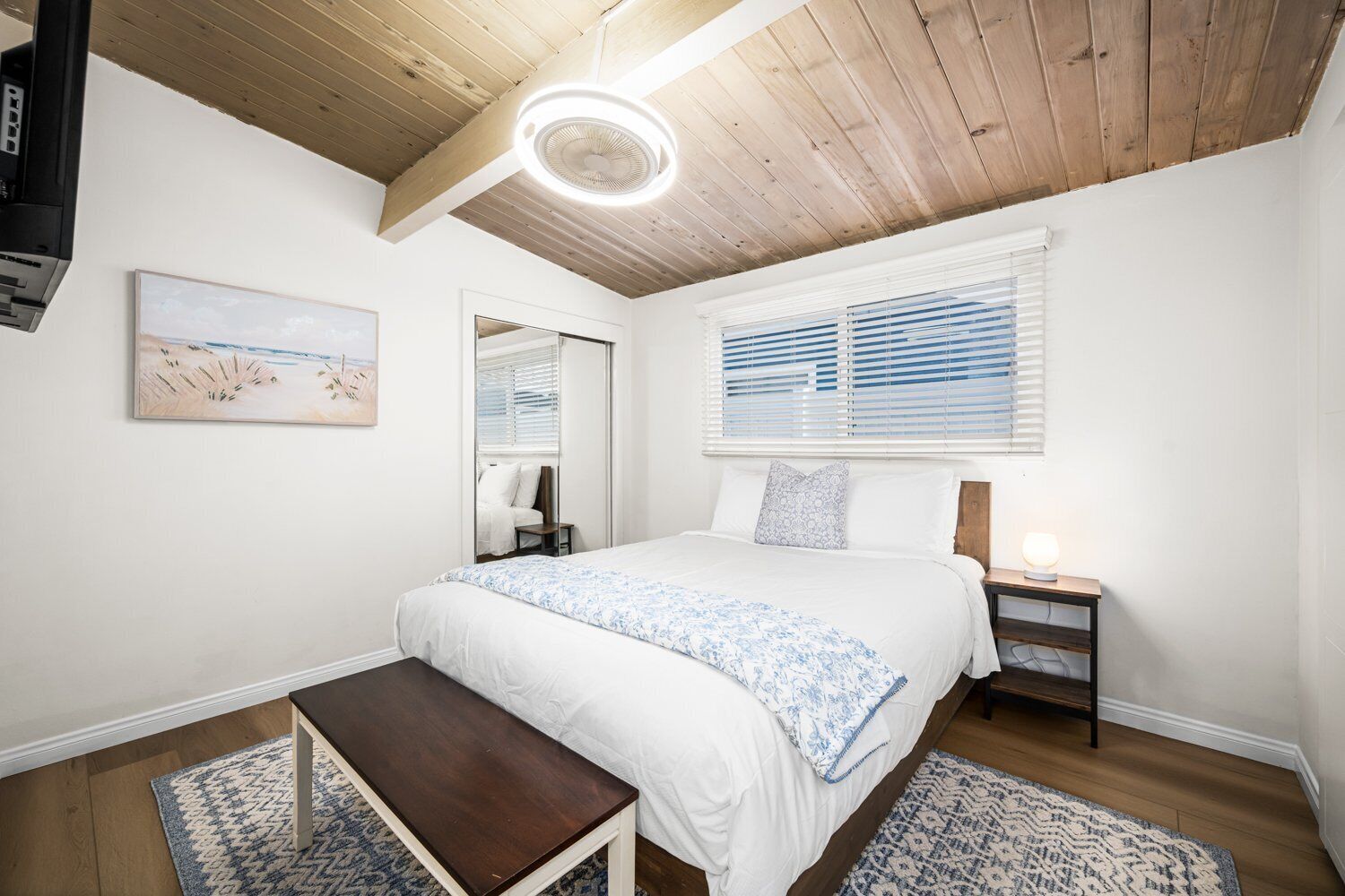 A bedroom with a large bed, a bedside table, a bench, a ceiling fan, and a window with blinds.