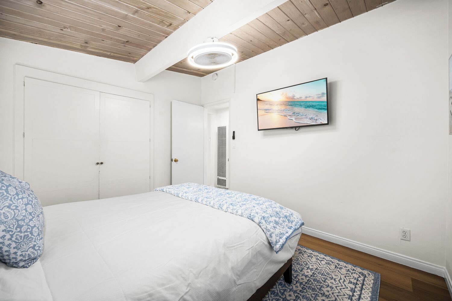 A bedroom with a bed, a ceiling fan, a TV on the wall, and a patterned rug.
