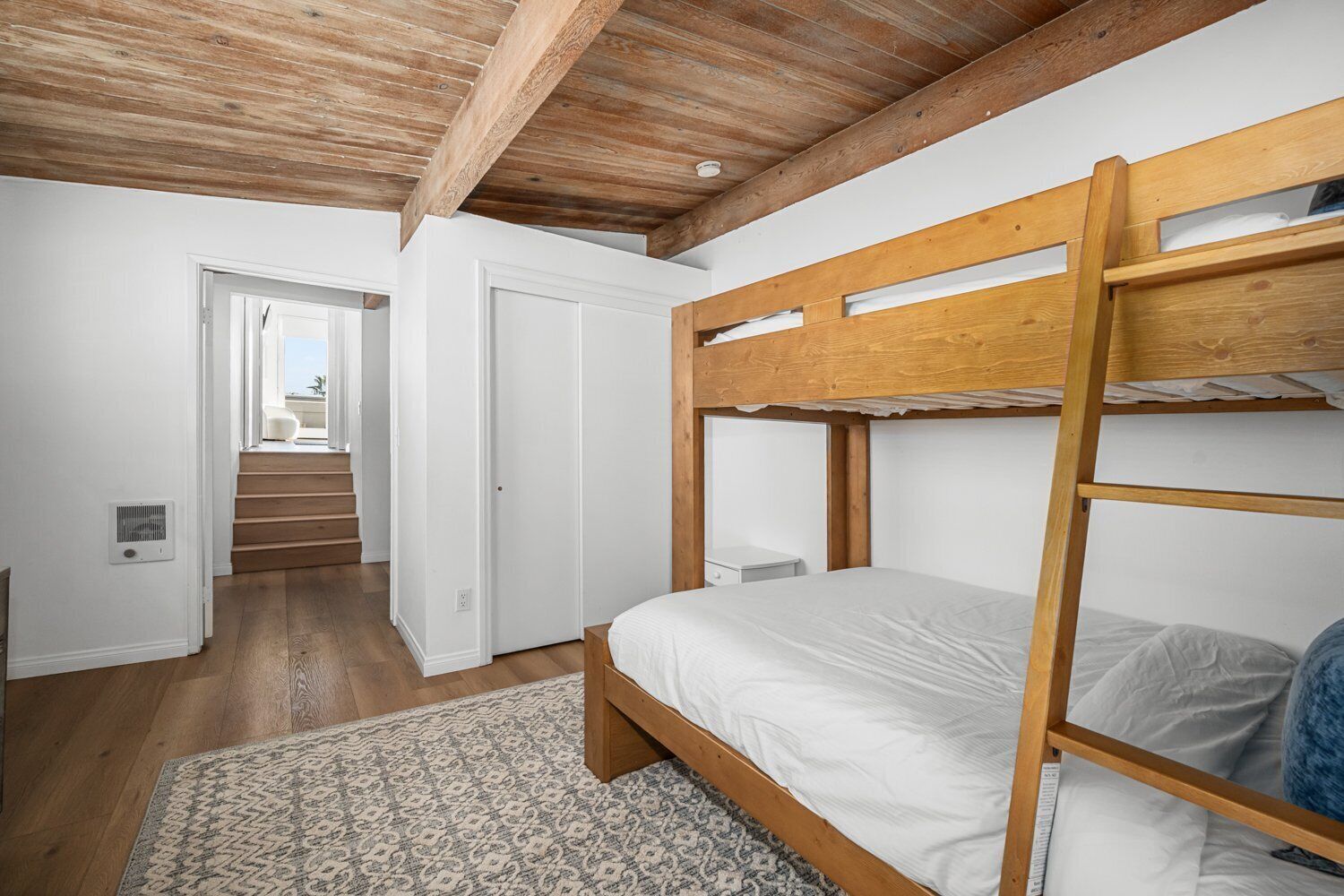 Een slaapkamer met een stapelbed, houten balken in het plafond, een kamerkleed met een patroon en uitzicht op een trap die naar een balkon leidt.