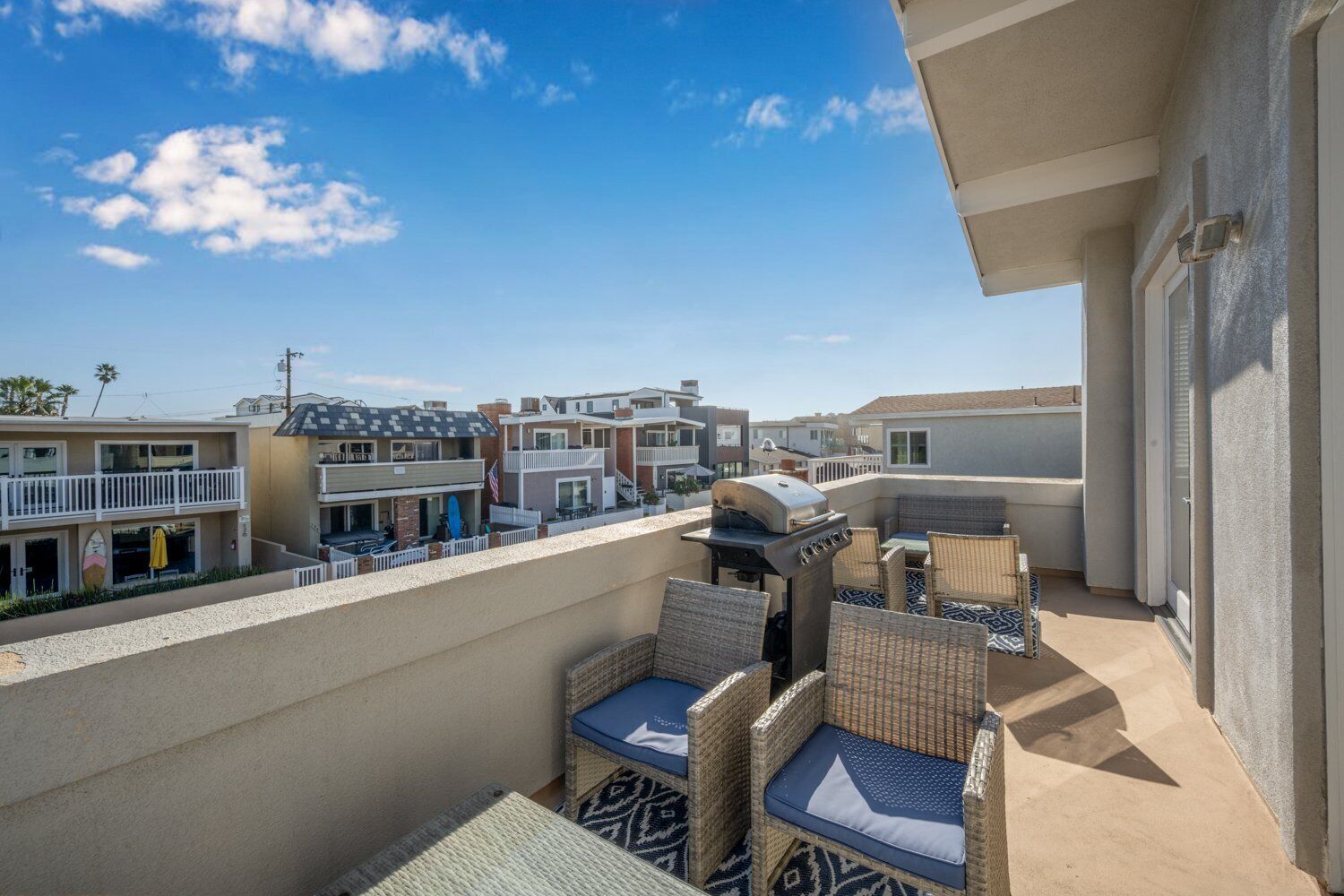 Terraza en la azotea con mobiliario de mimbre y una parrilla, con vistas a una zona residencial con casas y cielo despejado.