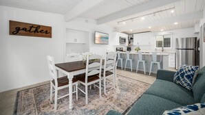 Una cocina moderna con armarios blancos, una zona de comedor con mesa de madera y sillas blancas, y un salón con sofá verde.