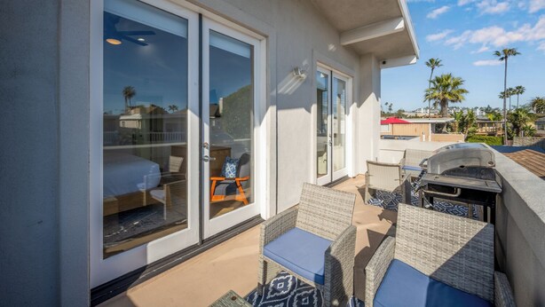 Un balcón con mobiliario de mimbre y una parrilla, puertas de vidrio que dan a un dormitorio, y vistas a palmeras y casas.