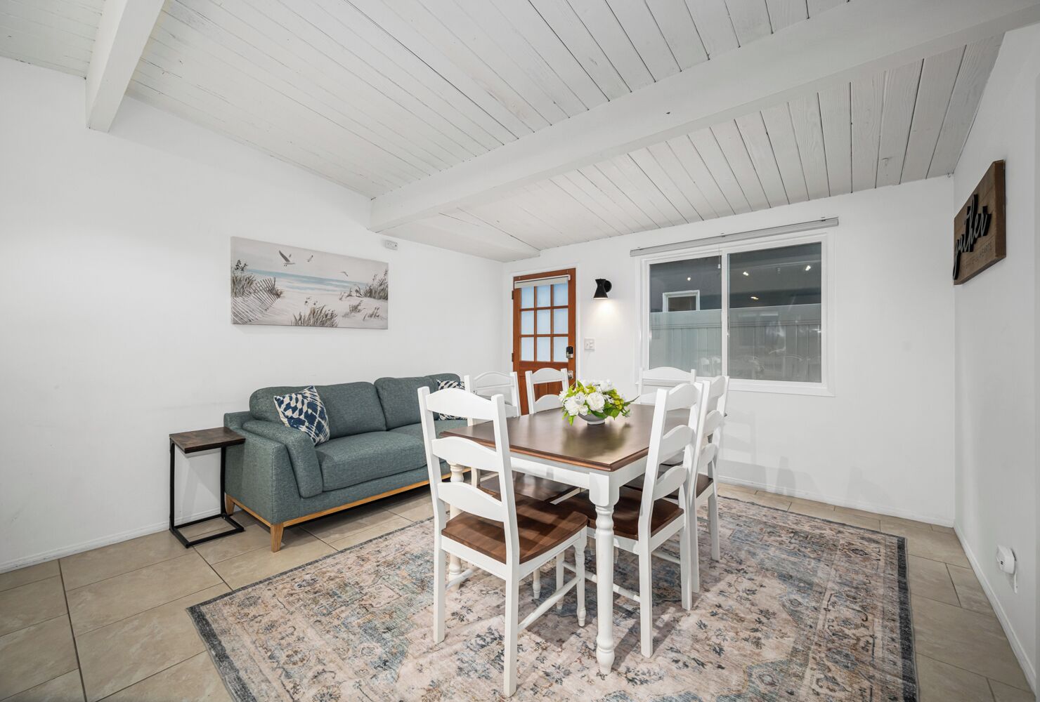 A dining area with a wooden table and white chairs, a grey sofa, and a patterned rug.