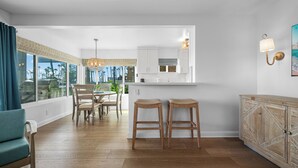 A modern kitchen with a dining area, wooden flooring, and large windows offering a view of the outdoors.