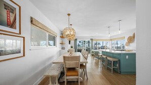 A spacious kitchen with a wooden dining table, a bar area, and large windows offering a view of palm trees.