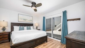 Un dormitorio con una cama, dos mesitas de noche, un ventilador de techo, un ventanal con cortinas y vistas a una escalera.