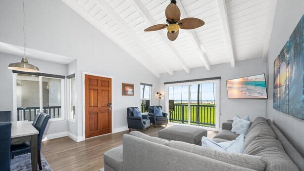 A spacious living area with a large gray sofa, a dining table with chairs, a ceiling fan, and a view of the beach through a sliding glass door.