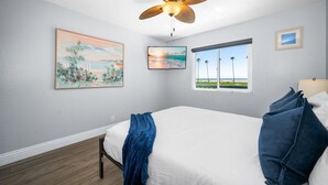 A bedroom with a bed, a ceiling fan, two windows with a view of the beach, and two framed pictures on the wall.