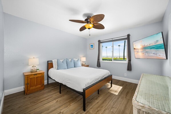Une chambre à coucher comprenant un lit, une table de chevet, une lampe, un ventilateur de plafond, une fenêtre donnant sur la plage et un fauteuil en osier.