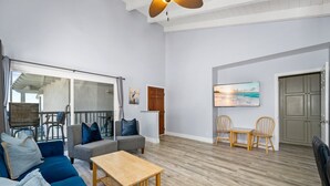 A spacious living room with a blue sofa, a wooden coffee table, and two wooden chairs. A large window offers a view of the outdoors, and a painting is on the wall. - Balboa Peninsula Hidden Gem with Private Balcony & Stunning Views (Newport Beach)