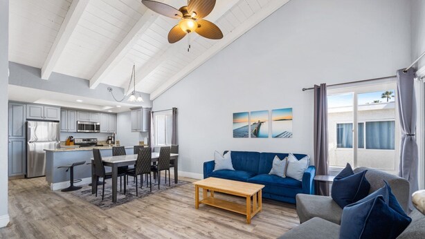 A modern living room with a blue sofa, a wooden coffee table, and a dining area with a kitchen in the background. - Balboa Peninsula Hidden Gem with Private Balcony & Stunning Views (Newport Beach)