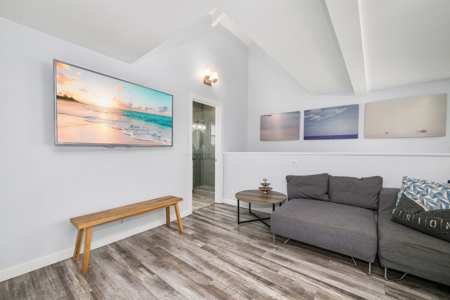 Een moderne woonkamer met een grijze bank, een houten zitbank en een groot schilderij met een strandthema aan de muur.