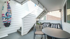 A covered outdoor seating area with a table and chairs, a surfboard, and string lights. - Perfect Surf Casa in Newport Near Beach and Pier (Newport Beach)